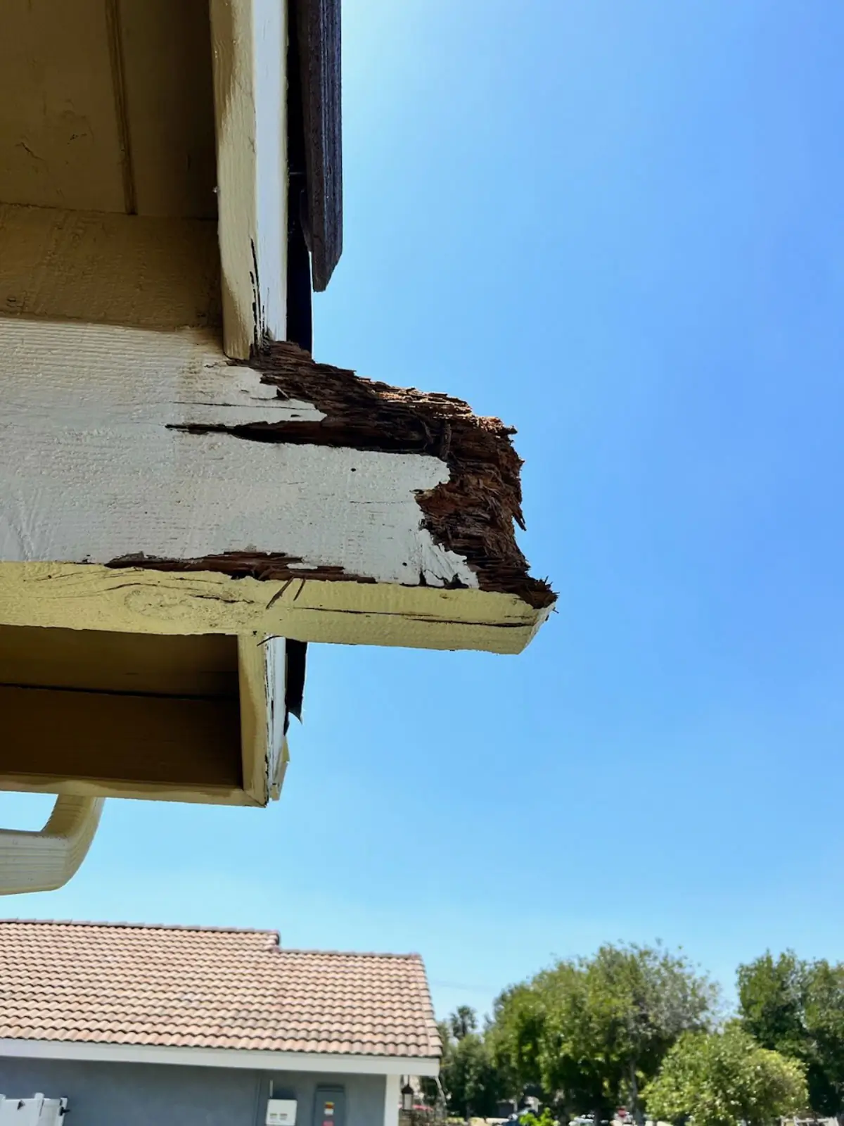 Image of a damaged wooden eave on a residential house with significant rot and deterioration. The eave is part of the exterior structure under the roof, showing exposed wood fibers and missing chunks due to decay.