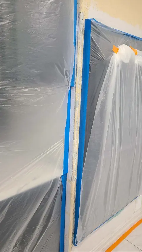 Close-up of a room corner covered in plastic sheeting with blue painter's tape and orange masking tape visible.