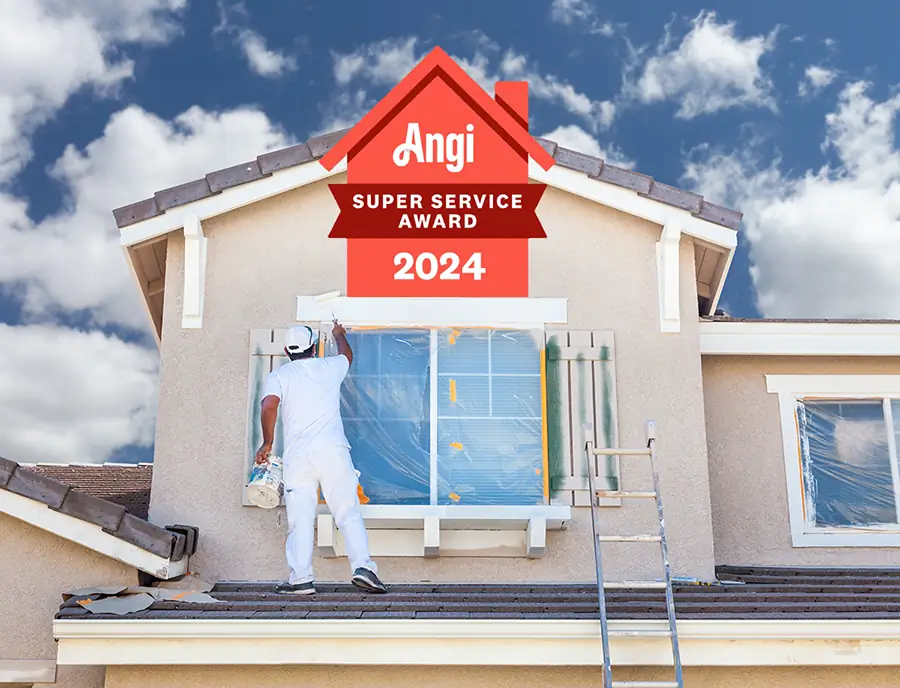 Professional house painter in white uniform painting an exterior window of a beige house with a blue sky background, featuring the 2024 Angi Super Service Award badge on top.