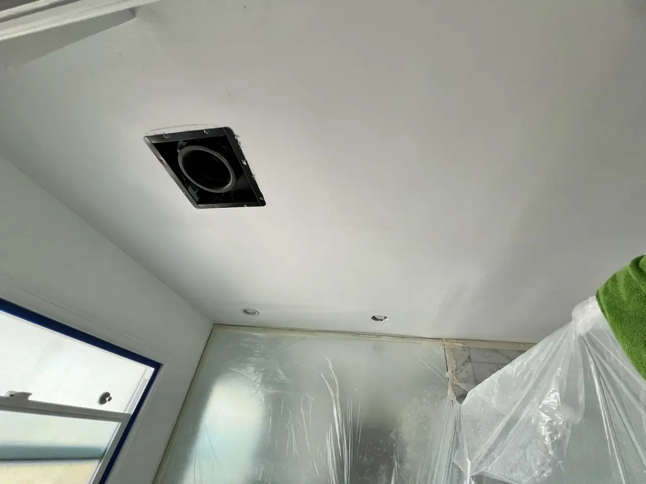 Bathroom wall in Costa Mesa before renovation, featuring a window, covered toilet, and sink area prepared with protective sheets